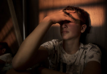 Teenage boy sitting alone, hand over his eyes, in the fading light.